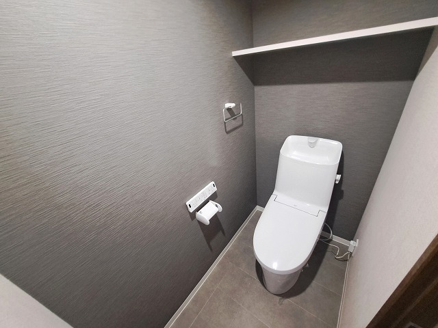 グレーのクロスと床材で仕上げられた、落ち着きのあるトイレ空間。シンプルながらもスタイリッシュな印象で、清潔感とデザイン性を兼ね備えています。上部には便利な棚付きで、トイレットペーパーや掃除用品の収納に重宝します。コンパクトながらも圧迫感のない設計で、毎日使う場所だからこそ快適さを追求。来客時にも気兼ねなく使っていただける、上質な雰囲気が魅力です。生活感を抑えた空間で、日常にちょっとした上質さをプラスしてくれるトイレです。